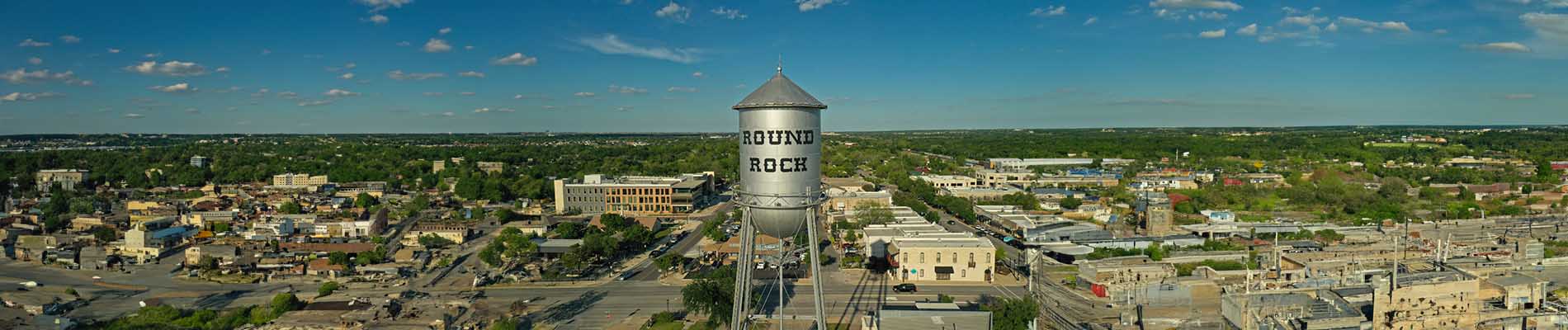 The image shows a clear daytime sky with a prominent water tower in the background, situated in a suburban area with houses, trees, and a wide-angle perspective that includes part of a roadway.