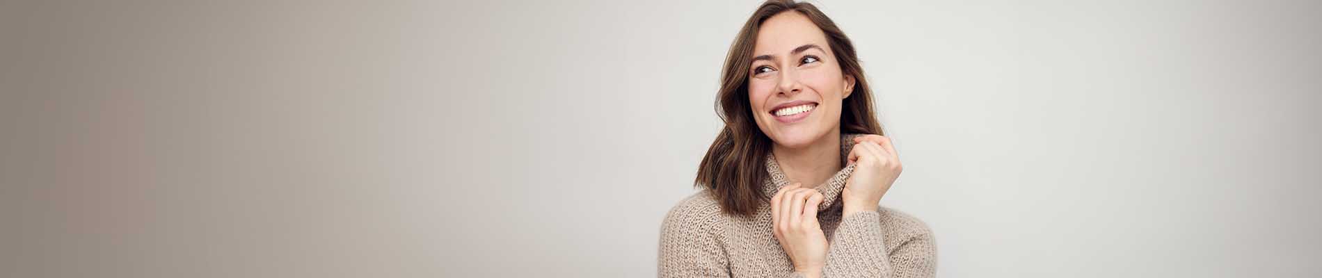 The image is a photograph of a woman with light skin, smiling at the camera. She appears to be in her late twenties or early thirties and has long hair. Her eyes are looking directly at the camera, and she is holding up her index finger near her mouth as if she s making a point or emphasizing something. The background is plain and light-colored, which suggests that this could be a stock photo used for various purposes such as advertising, personal branding, or lifestyle content.