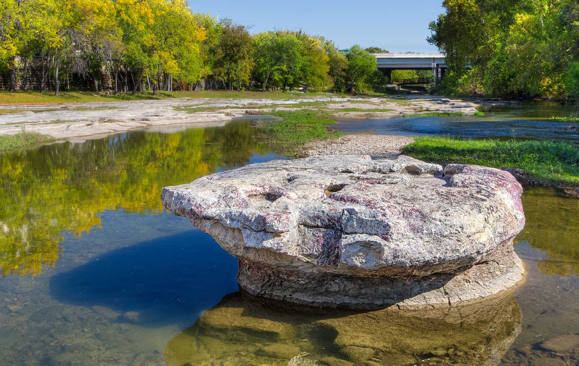 The image shows a natural landscape with a large rock formation prominently situated on a riverbank, surrounded by water and bordered by a forested area with autumnal foliage.